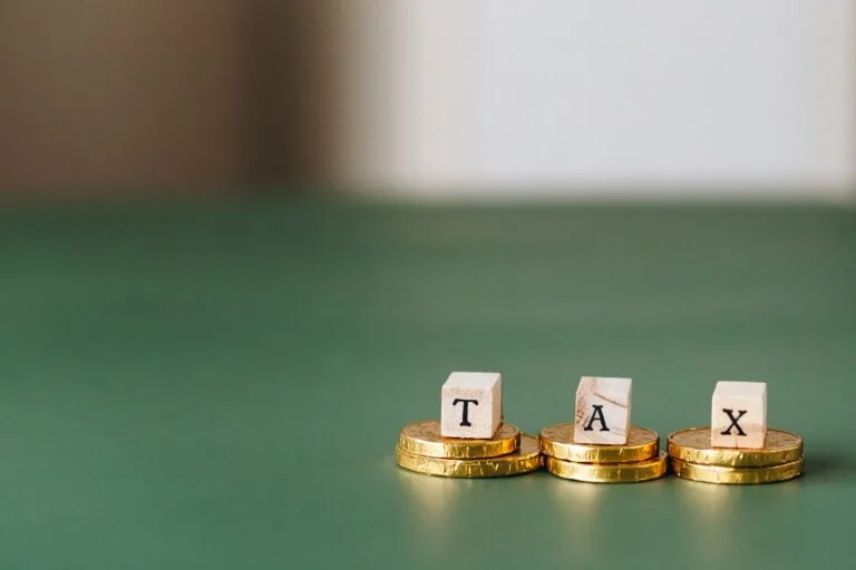 Compensação tributária: como usar créditos para quitar débitos com o fisco 2 Stacked gold coins with wooden blocks spelling TAX on a green backdrop.
