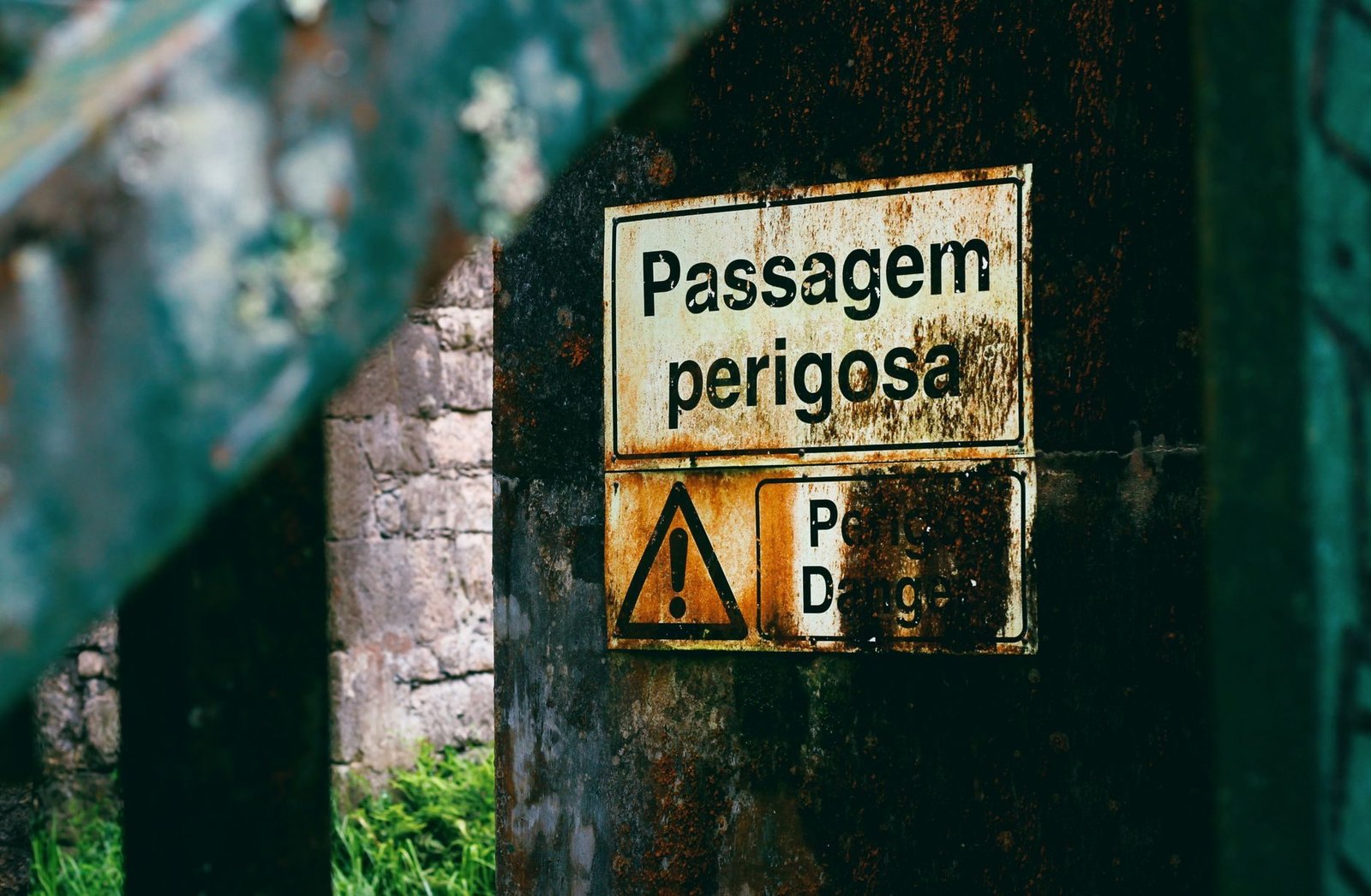 Old rusty warning sign in Portuguese amidst urban decay and vegetation.