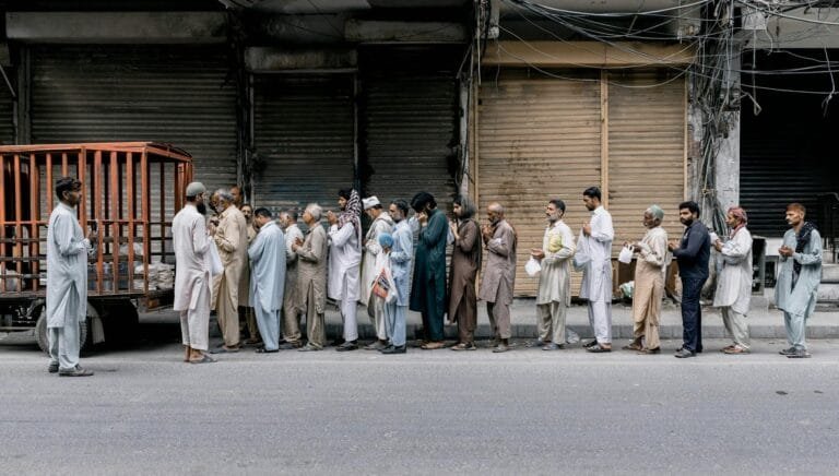 A group of adults standing in line for charity aid on a city street in Faisalabad, Pakistan.