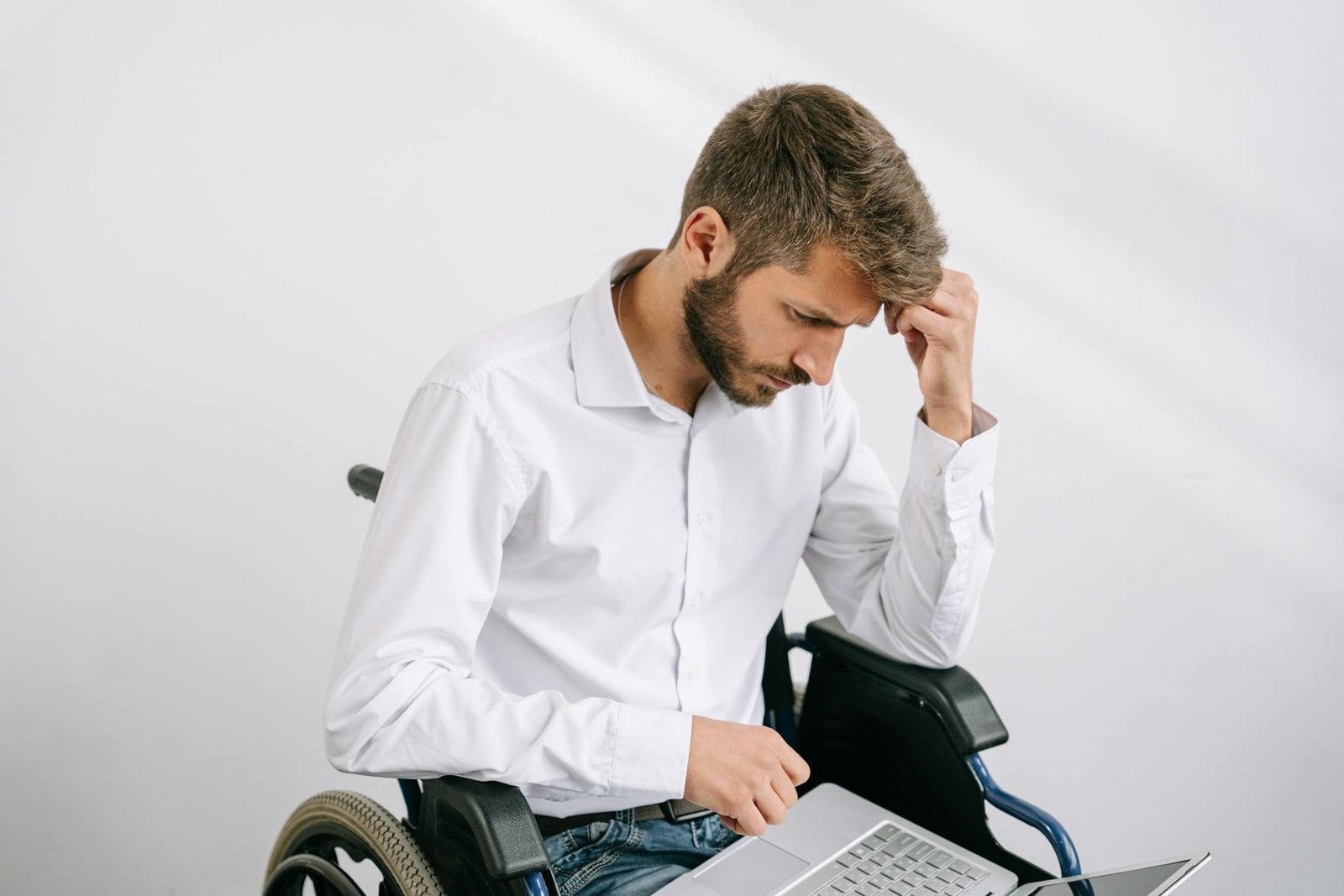 A young bearded man in a wheelchair works on his laptop indoors, wearing a white