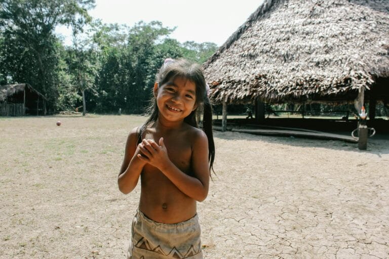 A young girl smiling in a rural village setting in Iquitos, Peru, with traditional huts in the background.