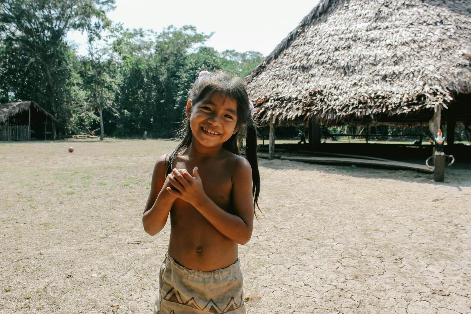 A young girl smiling in a rural village setting in Iquitos, Peru, with traditional huts in the background.
