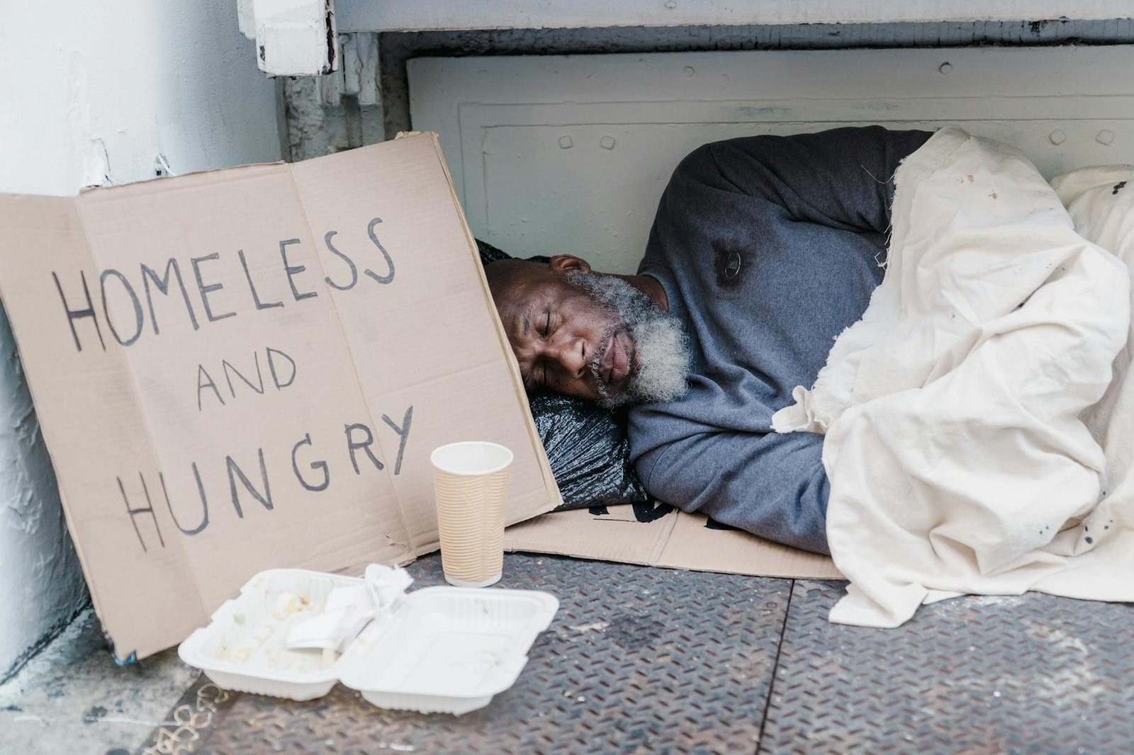 Elderly homeless man sleeping on the street with a sign reading 'Homeless and Hungry'.