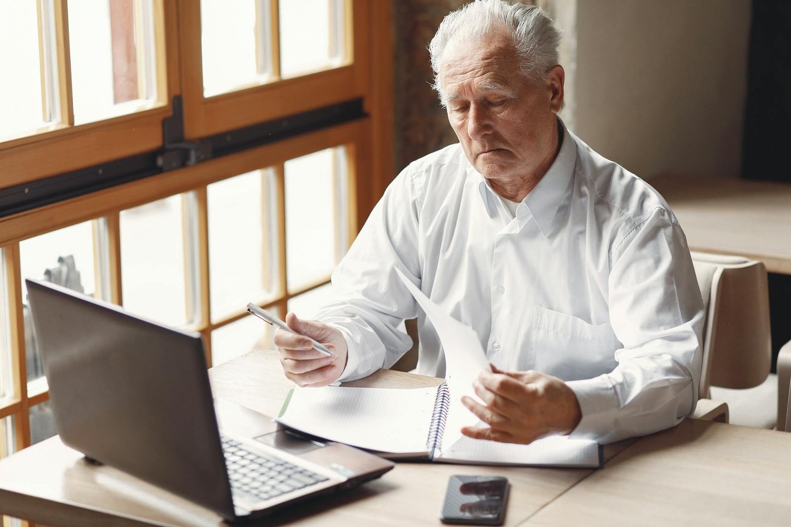 Serious gray haired aged man in white shirt sitting at table and browsing laptop
