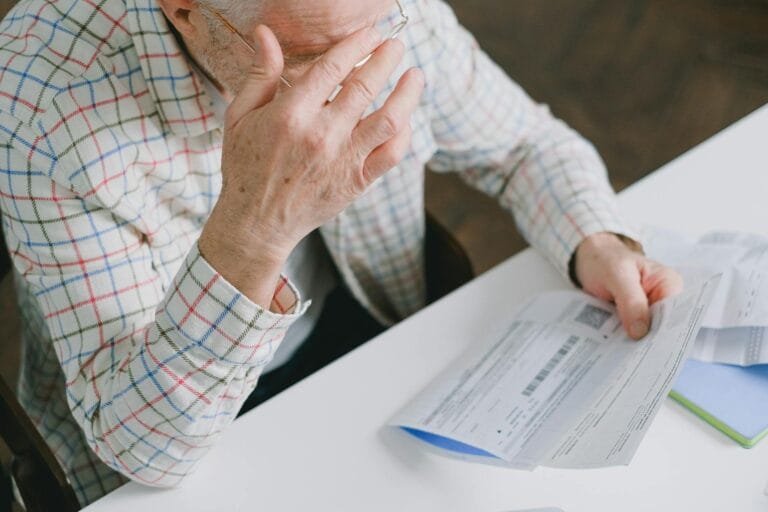 Elderly man in patterned shirt reading and holding bills at a home table, appear