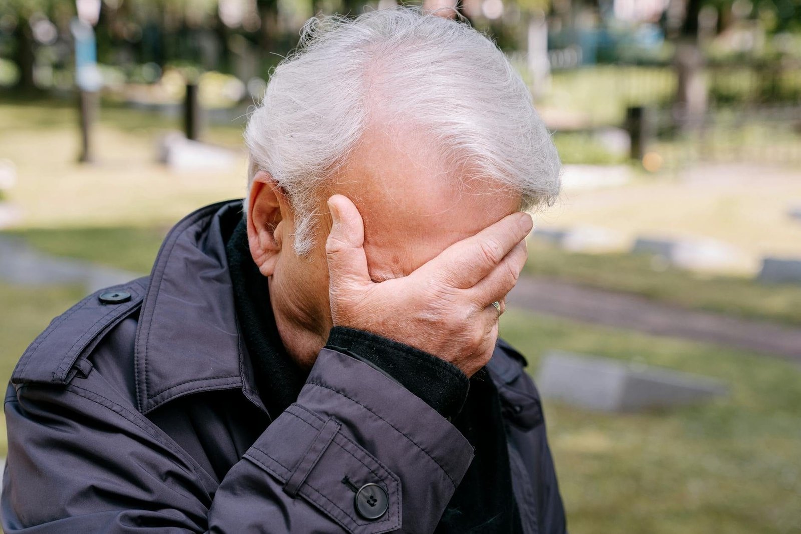 Senior adult man in grief, covering face with hand, in a cemetery setting.