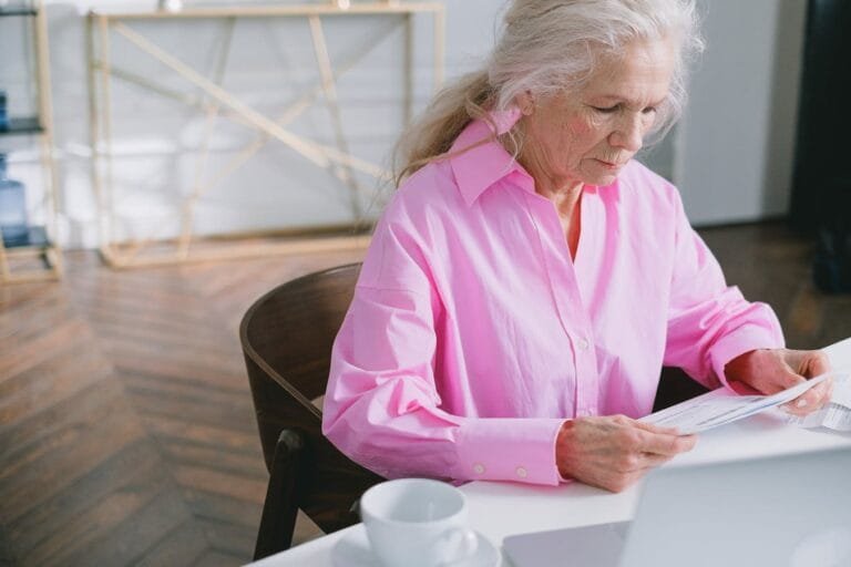 Elderly woman in pink shirt reading papers at home office desk, focused on work.