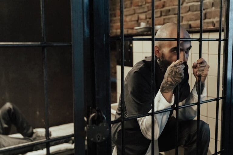 Bald man gripping prison bars inside a cell, displaying tattoos and wearing a uniform.