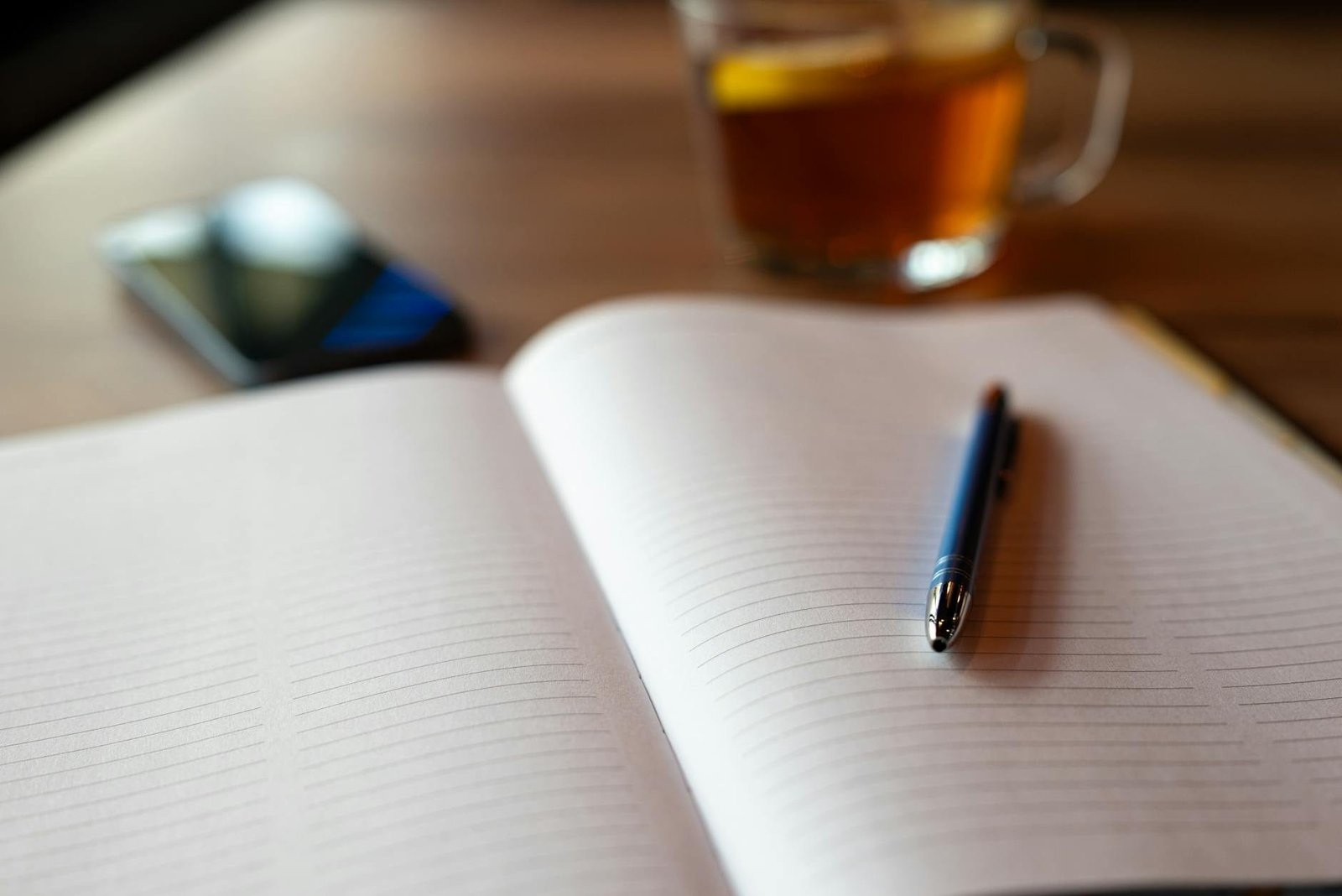 An open notebook with a pen, smartphone, and tea on a wooden table. Perfect for