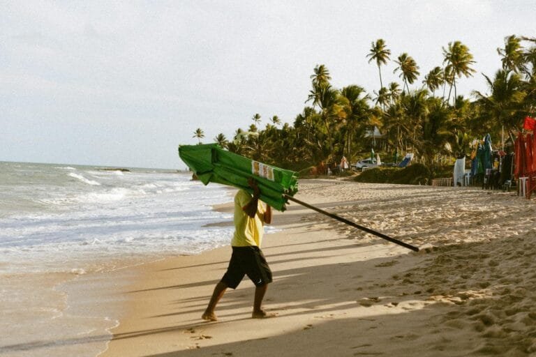 A beach vendor carrying an umbrella along a tropical shoreline in João Pessoa, B