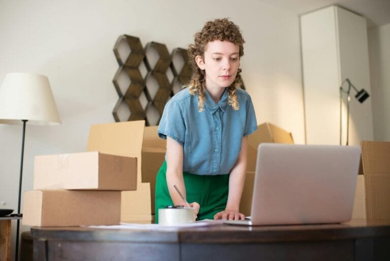 A young woman working on her laptop surrounded by cardboard boxes, indicating on