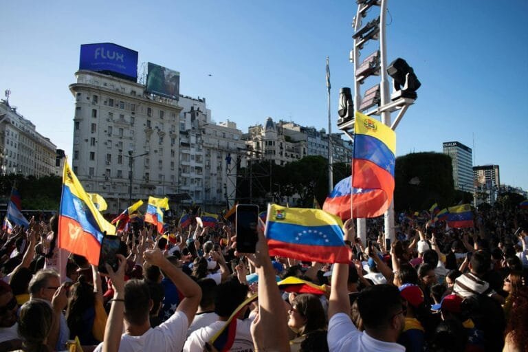 A large crowd waves Venezuelan flags during a protest in a bustling city square.