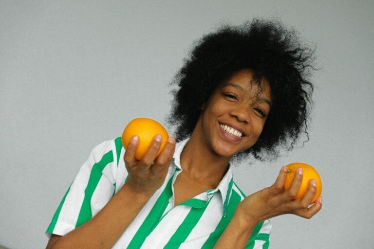 Cheerful woman with afro hair holding oranges indoors, radiating joy and vitalit