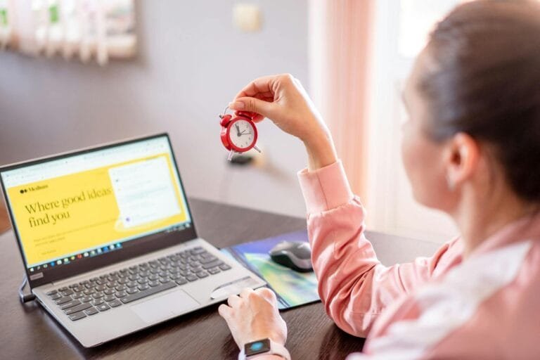 PPP e LTCAT: o que são e por que esses documentos definem o reconhecimento do tempo especial 5 Woman holding a small clock at a desk with an open laptop screen displayed.