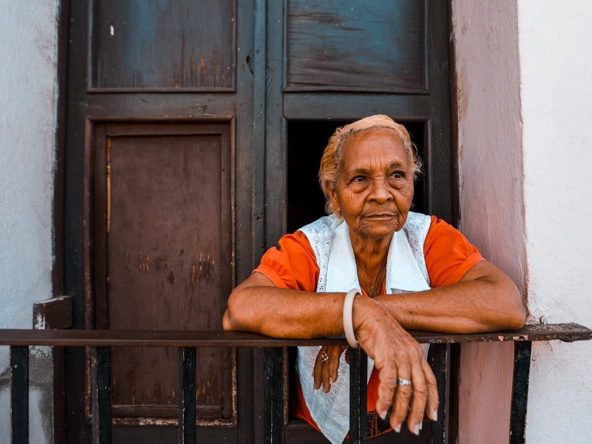 Pensão por Morte: Beneficiários e Ordem de Habilitação 1 Senior woman in orange shirt leaning on a metal railing balcony.