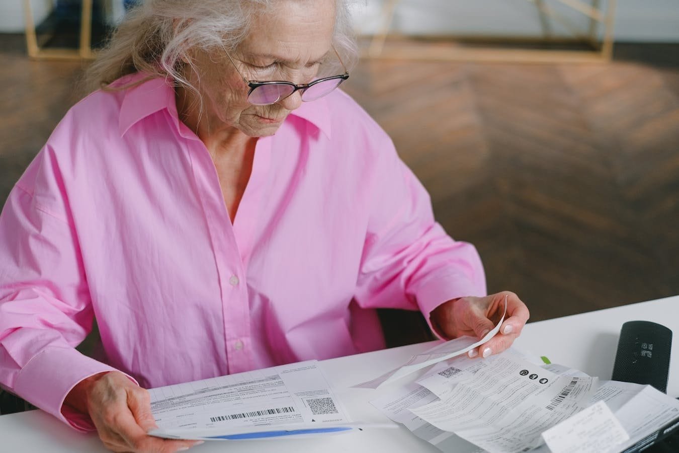 Elderly woman in pink blouse reading documents at a table indoors.