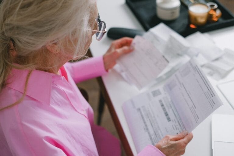 Revisão do BPC em 2026: quem pode ser convocado pelo INSS 5 An elderly woman in glasses holds and reads important papers at a table indoors.