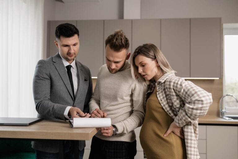 A young couple reviews and signs real estate documents with an agent in a modern kitchen setting.