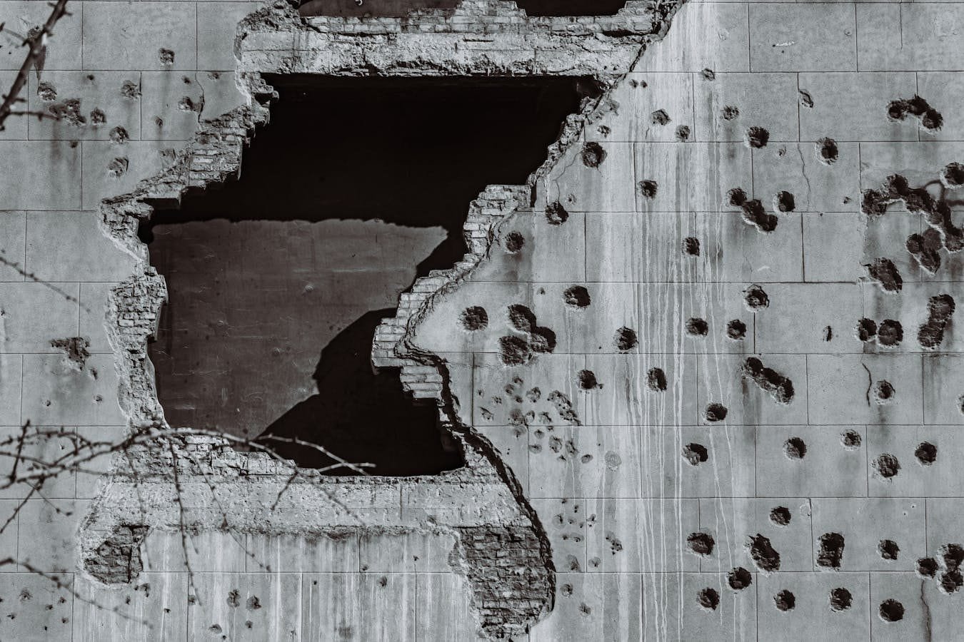 A dynamic black and white photo of a damaged concrete wall, featuring a large hole and multiple bullet impact marks, sym
