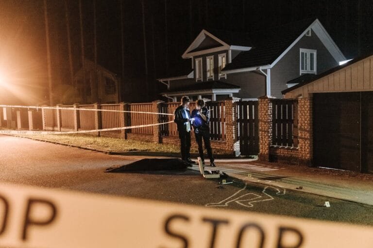 Police officers investigating a nighttime crime scene outside a residential house with barrier tape and evidence markers