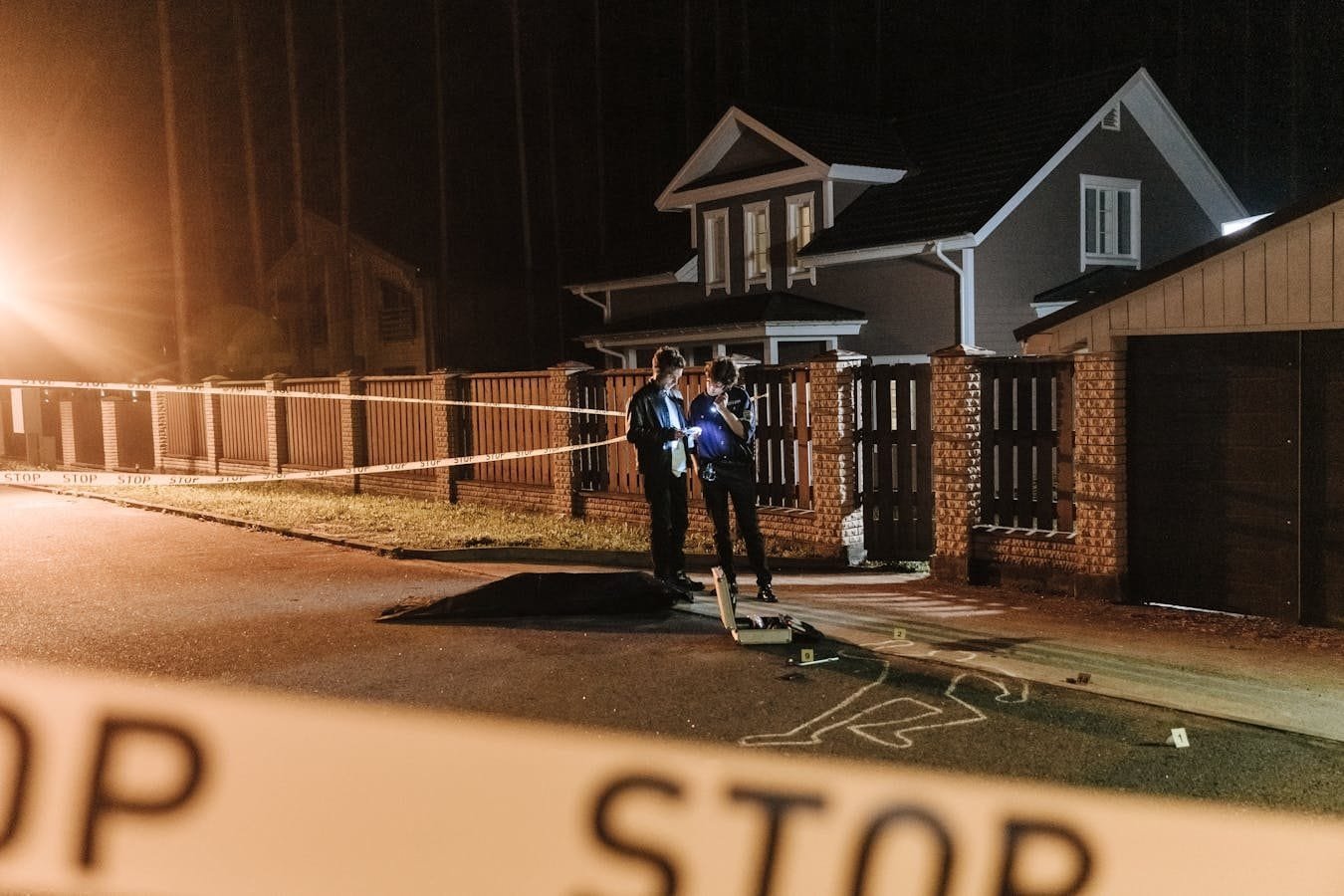 Police officers investigating a nighttime crime scene outside a residential house with barrier tape and evidence markers