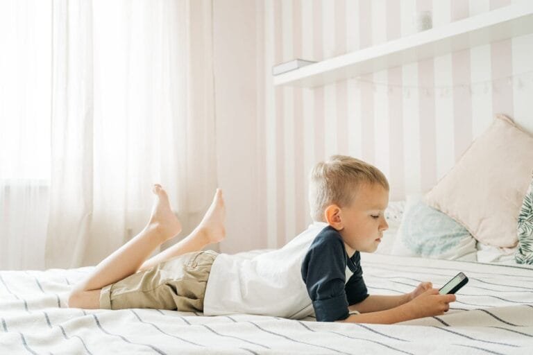 Young boy lying on bed using smartphone in a bright room, enjoying leisure time.