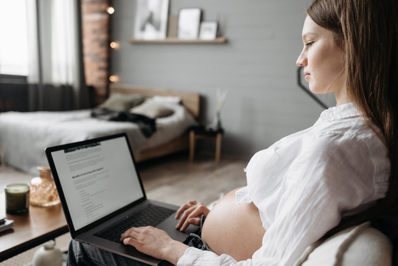 Pregnant woman working on a laptop in a cozy, light-filled bedroom. Side view of her baby bump.