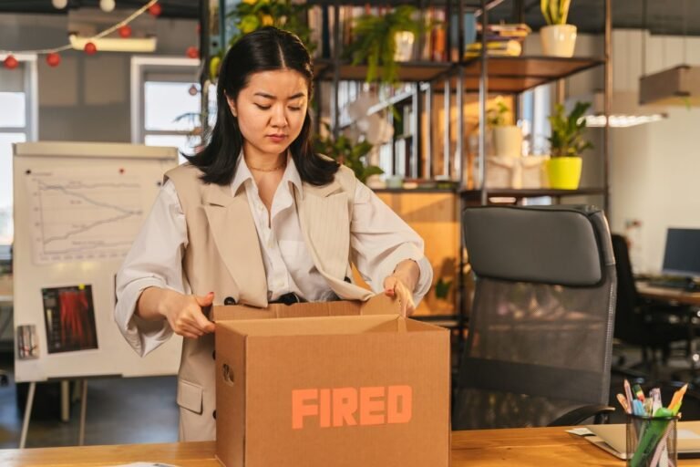An Asian woman packs a box labeled FIRED, symbolizing job loss in an office setting.