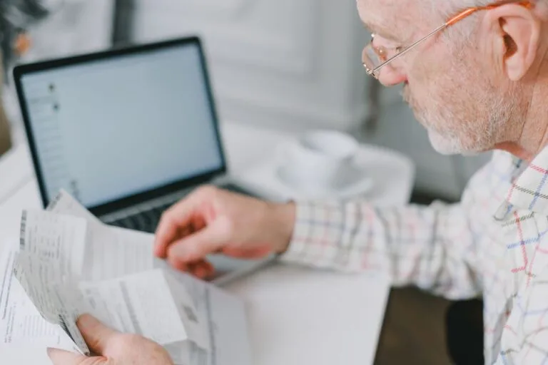 Acumulo de pensão por morte com aposentadoria: o que mudou e o que permanece 5 Elderly man wearing eyeglasses reading documents next to a laptop at home.