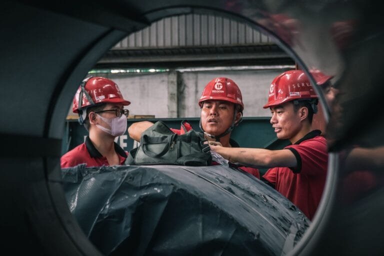 Agentes Nocivos para Aposentadoria Especial em 2026 7 Three workers in safety helmets collaborate inside a factory setting, emphasizing teamwork and safety.