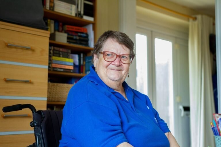 Senior woman in blue shirt sitting indoors with a smile and bookshelves background.