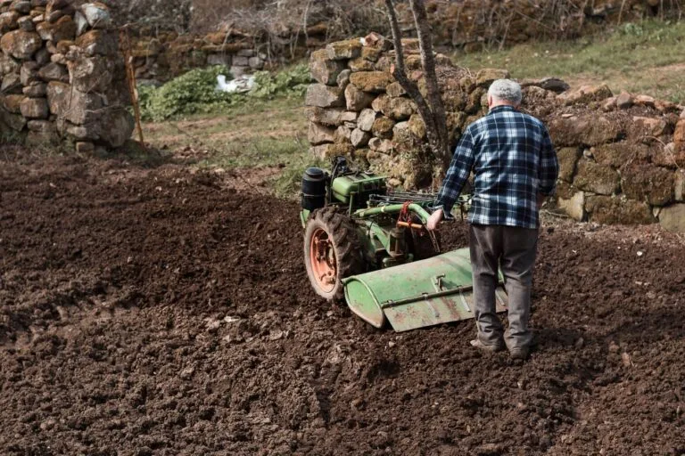 Elderly man using a green tiller in a rustic field with stone walls, embodying traditional farming.