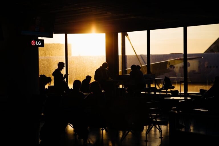 Atraso e cancelamento de voo: seus direitos a assistência e indenização 6 Travelers sitting in an airport terminal at sunset, silhouetted against the backdrop of an airplane.