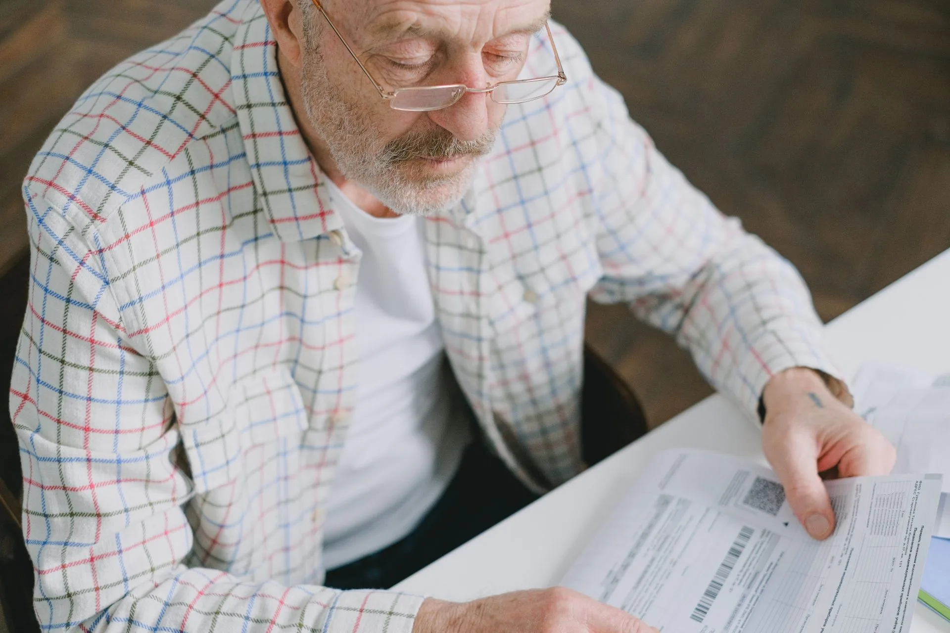 Elderly man in glasses carefully examines documents at home. Thoughtful and focused.