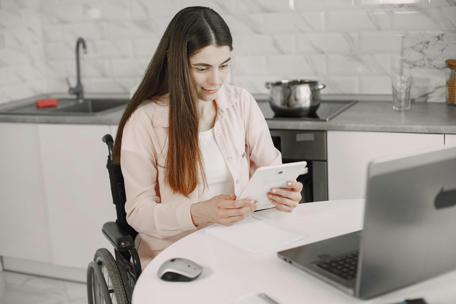 Woman in wheelchair engaged with tablet at kitchen table, creating an inclusive work environment.