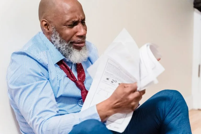African American man in distress with paperwork, sitting against a wall indoors.