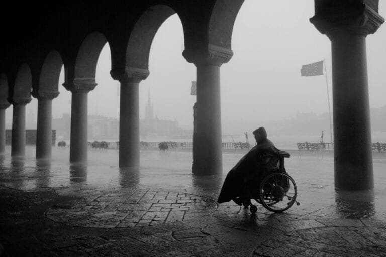Black and white photo of a person in a wheelchair under archways on a rainy day in Stockholm.