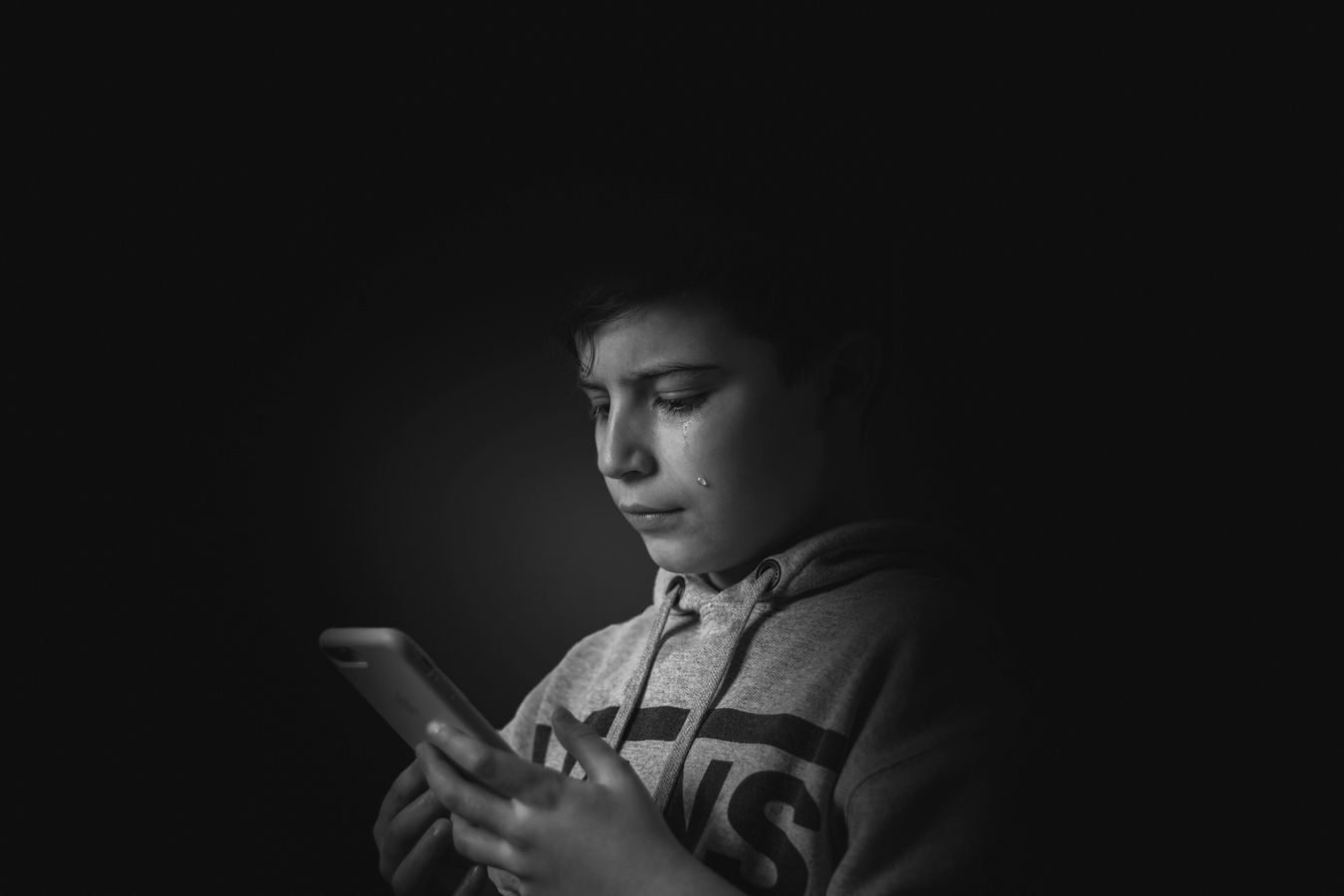 Ciberbullying contra criancas e adolescentes: o que a familia e a escola podem fazer 1 A monochromatic portrait of a boy holding a smartphone, expressing sadness.