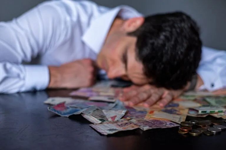 Cobrança vexatória de dívidas pode gerar indenização e crime 3 A man in a white shirt rests his head on a table covered with Argentine pesos and coins.