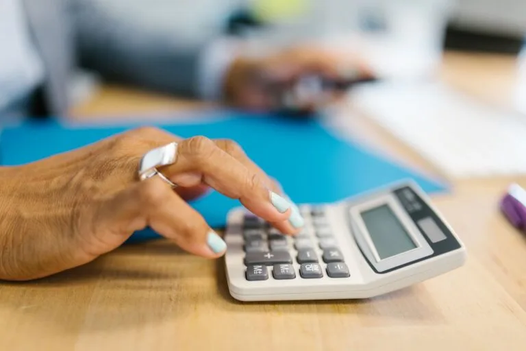 Manicured hand using a calculator on a desk with a blue folder, shallow depth of field.