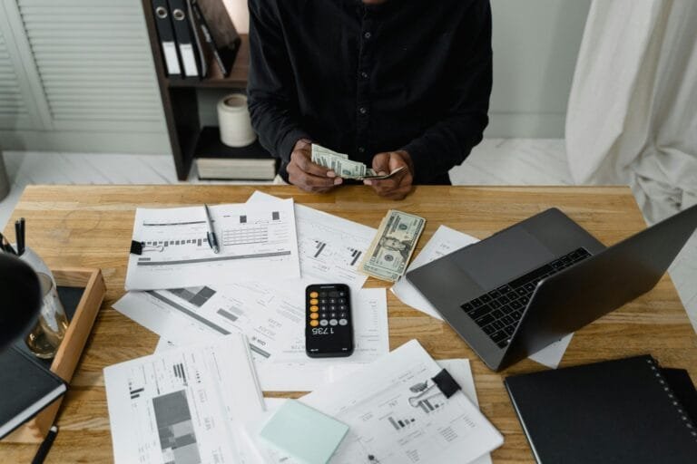 Complementação de Contribuição Previdenciária: Quando Pagar 5 A person counts dollar bills at a desk with a laptop, papers, and calculator.