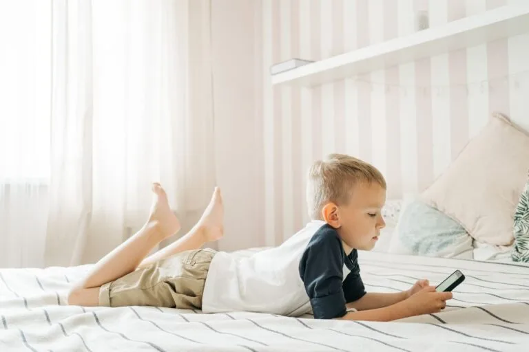 Young boy lying on bed using smartphone in a bright room, enjoying leisure time.