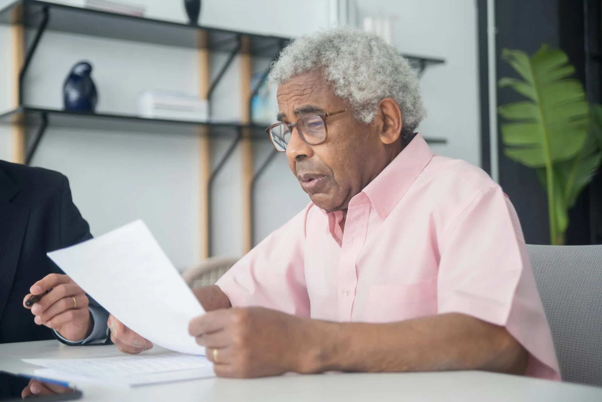 Elderly man with glasses reading a document in a home office setting, reflecting focus and concentration.
