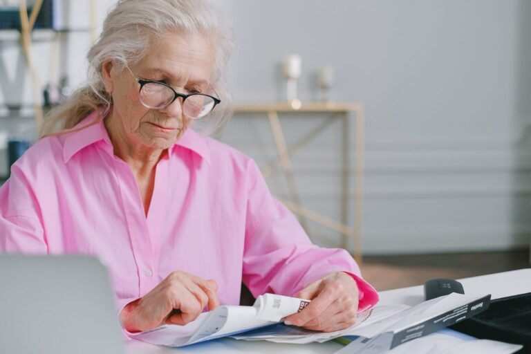 Elderly woman in pink shirt reading documents at a desk indoors.