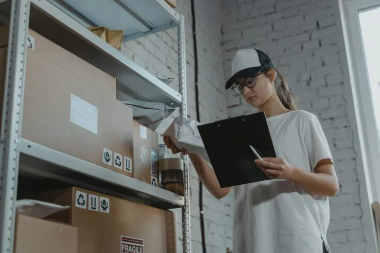 DIFAL do ICMS: Impactos no E-commerce 7 Warehouse worker organizing packages on shelf with clipboard.