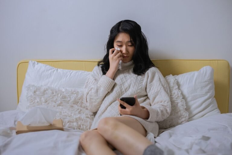 Asian woman in a sweater, looking sad and wiping tears, holding a smartphone seated on a b