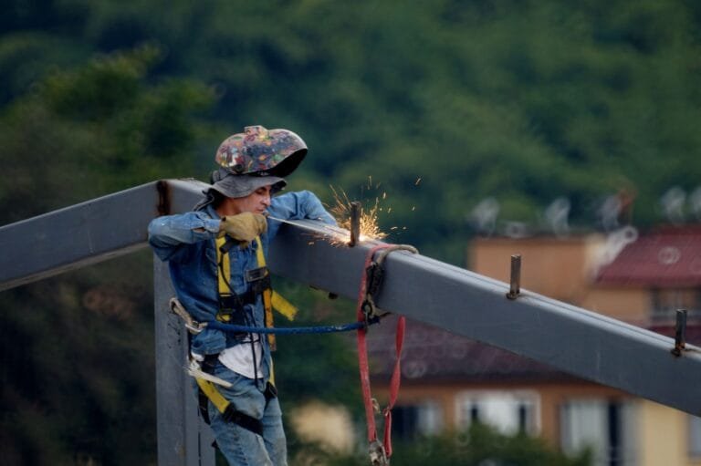 EPI e Aposentadoria Especial 2026: Uso Elimina o Direito? 2 Construction worker welding steel beam on site, wearing safety gear and helmet.