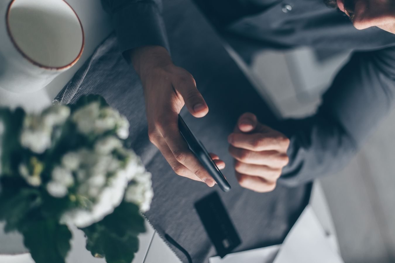Top view of a mans hands holding a smartphone indoors with a cup and flowers nearby.