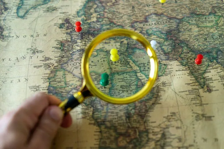 Close-up of a hand holding a magnifying glass over a world map with colorful pins indicati
