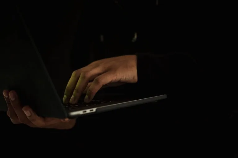 Crop anonymous male wearing black hoodie working on modern netbook while standing in dark 